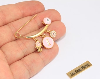 Puede incluir: Alfiler de seguridad chapado en oro con detalles de esmalte rosa y colgantes. Los colgantes incluyen una mano de Hamsa, un círculo rosa con la letra "I" y un dije en forma de ojo. El alfiler se sostiene en una mano.