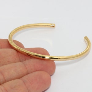 Peut inclure: Un bracelet manchette ouvert de couleur or. Le bracelet a un design simple et arrondi et est fait d'un matériau métallique brillant. Le bracelet est présenté sur un fond blanc.