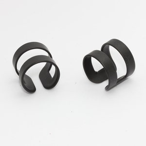 以下が含まれることがあります： ダブルループデザインの黒いイヤーカフ2つ。 イヤーカフはマットブラック素材で、白い背景に表示されています。 モダンなジュエリーアクセサリーです。