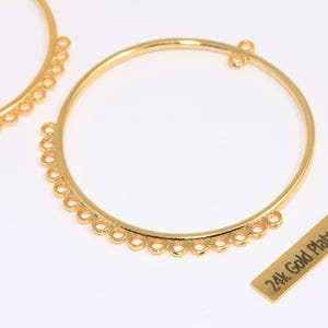 以下が含まれることがあります： 下部に一連の小さな穴がある、2つの金色の円形イヤリングコンポーネント。「24k Gold Plated」の文字が小さな金の長方形にあります。 ジュエリー作りの材料。