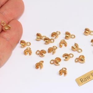 May include: A selection of small, gold-coloured crimp bead end caps with a loop for jewellery making. The caps are scattered on a white surface, with a small brass rectangle that reads "Raw Brass".