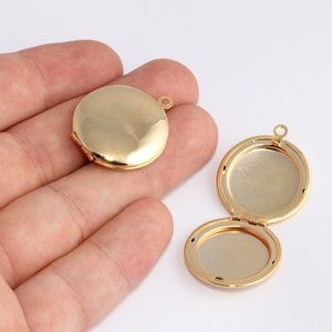 Puede incluir: Dos medallones redondos de color dorado. Un medallón está cerrado y el otro está abierto, revelando el interior. Ambos medallones tienen un pequeño lazo en la parte superior para sujetarlos a una cadena. Los medallones están sobre un fondo blanco.