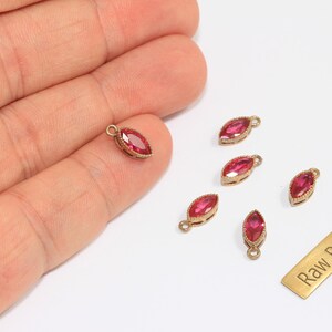 May include: Marquise-shaped red gemstone charms with gold-colored metal settings. The charms are small, with a loop at the top for attaching to jewelry. A small brass tag with the text "Raw Brass" is also present.