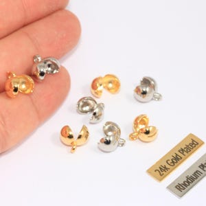 May include: Close-up of several gold and silver-colored jewelry findings. The findings are small, rounded, and have a clasp design. Two rectangular tags read "24k Gold Plated" and "Rhodium Plated".