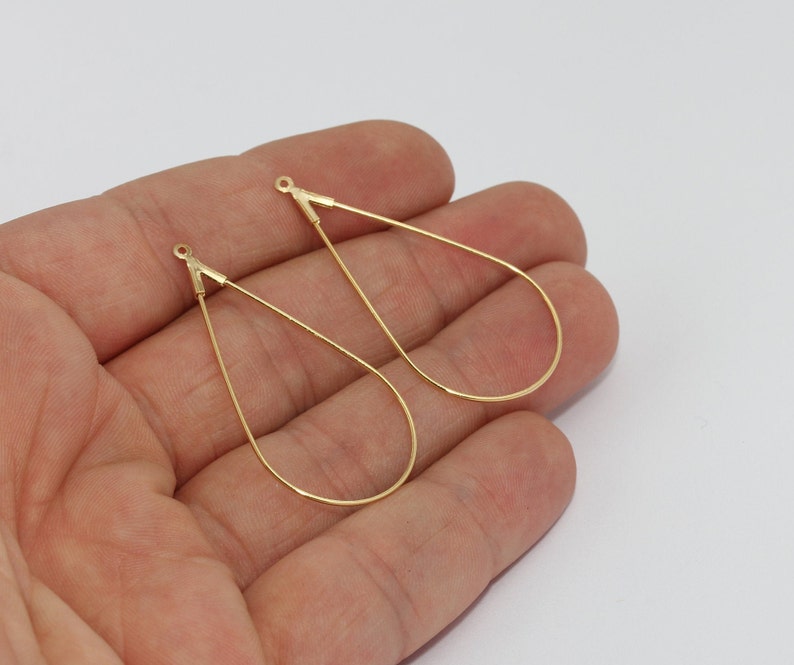 May include: Two gold-coloured teardrop-shaped earring components. Each earring has a small loop at the top and a slender, curved wire forming the teardrop shape. The earrings are displayed against a white background.