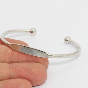 May include: A silver-colored cuff bracelet with a rectangular center and rounded ball ends. The bracelet is a simple, elegant design, suitable for everyday wear. The metal has a polished finish.
