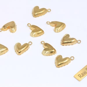 May include: A collection of gold-colored heart-shaped charms with small loops for attaching to jewelry. The charms are scattered on a white surface, with a small rectangular tag that reads "Raw Brass".