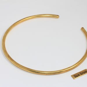 May include: A gold-colored, open-ended choker necklace. The necklace is a simple, curved design. A small, rectangular tag next to the necklace reads "Raw Brass".