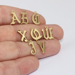 May include: A selection of gold-toned metal letter charms in an old English font. The letters A, B, C, X, Q, W, J, and V are shown. Each charm has a small loop at the top for attaching to jewellery or other crafts.