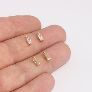 以下が含まれることがあります： 中央に透明な長方形の石が入った、4つの小さな金色の長方形のチャーム。各チャームの上部には、ジュエリーに取り付けるための小さなループが付いています。チャームは明るい背景に表示されています。