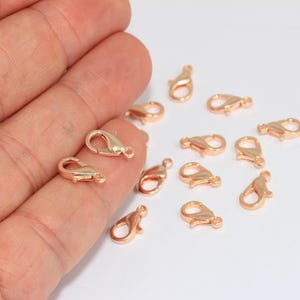May include: A collection of small rose gold lobster claw clasps for jewelry making. Each clasp features a spring-loaded mechanism and a small loop at the base for attachment. The metal has a smooth, polished finish with a distinct pinkish hue.