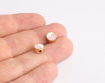 May include: Two gold-colored, round beads with a yin-yang design. The yin-yang symbol is in white and pink enamel. Each bead has a small hole for stringing. The beads are shown against a neutral background.