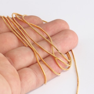 May include: A length of gold-colored chain, likely for jewelry making, is displayed against a plain white background. The chain has a delicate, textured appearance, and is held in a hand.