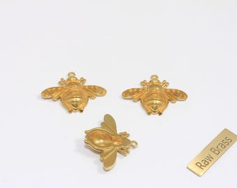 May include: Three gold-colored bee charms with detailed wings and body segments are displayed on a white background. One bee is upside down. A small gold rectangle with the text "Raw Brass" is also present. These charms are ideal for jewelry making.