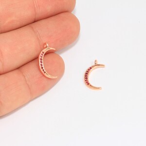 May include: Two rose gold crescent moon charms with red gemstones. The charms are small, with a loop at the top for hanging. One charm is held by a hand, while the other is on a white background. These charms are suitable for jewelry making.