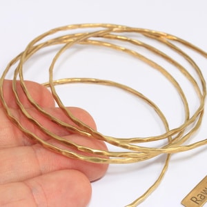 Op de afbeelding: Een verzameling gehamerde messing armbanden in een warme goudtint. De armbanden hebben een getextureerd, onregelmatig oppervlak. Een klein messing label met de tekst "Raw Brass" is ook zichtbaar.