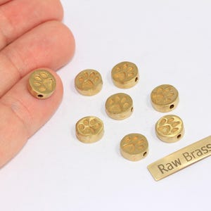 May include: A collection of small, round, brass beads with a paw print design. The beads are a warm gold color and have a small hole for stringing. A rectangular brass tag with the text "Raw Brass" is also present.
