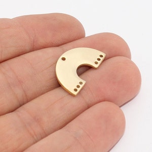 May include: A gold-colored, semi-circular metal jewelry finding with a rounded top and a curved cutout. It has three small holes on one end and three sets of three small holes on the other end. The finding is held in a hand.
