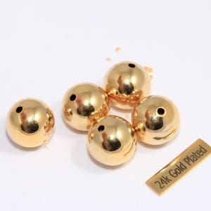 Könnte beinhalten: Fünf goldbeschichtete, runde Perlen mit glänzender Oberfläche, jede mit einem kleinen Loch. Ein kleines rechteckiges Etikett trägt die Aufschrift "24k Gold Plated". Die Perlen sind wahrscheinlich für die Schmuckherstellung.