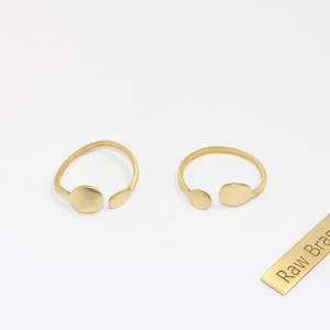 Könnte beinhalten: Zwei offene, goldfarbene Ringe mit minimalistischem Design, jeweils mit einem kreisförmigen Akzent. Ein kleines rechteckiges Etikett mit dem Text "Raw Brass" ist ebenfalls vorhanden. Die Ringe sind vor einem weißen Hintergrund platziert.