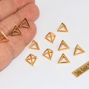 以下が含まれることがあります： オープンフレームの小さな3次元の金色のピラミッド型チャームのコレクション。黒で「24k Gold Plated」と印刷された長方形の金色のタグがあります。チャームは無地の白い表面に配置されています。