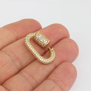 May include: A gold-colored, open carabiner clasp encrusted with small, clear crystals. The clasp is in an oval shape with a hinged top. The background is a plain, light color.