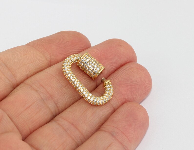 May include: A gold-colored, open carabiner clasp encrusted with small, clear crystals. The clasp is in an oval shape with a hinged top. The background is a plain, light color.