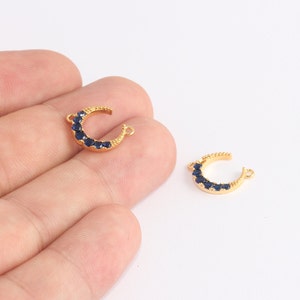 May include: Two gold-colored crescent moon charms with dark blue gemstones. The charms have small loops on each end for attaching to jewelry. The gemstones are set along the curve of the moon.