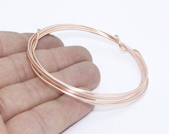 Puede incluir: Una bobina de alambre de color oro rosa, enrollada firmemente en forma circular. El alambre se sostiene en una mano sobre un fondo blanco. El alambre parece nuevo y sin usar, y probablemente sea para hacer joyas u otras manualidades.