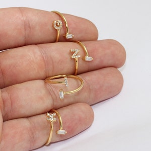 May include: Gold-toned adjustable rings with initial letter designs. Each ring features a different letter, adorned with small, clear gemstones. The rings are open-ended, with a gemstone accent on the opposite side of the initial.