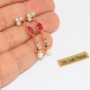May include: Gold-plated drop earrings with red, white, and blue gemstones and a pearl. The earrings have a floral design at the top and a teardrop-shaped red stone in the centre. The earrings are held in a hand. The text "24k Gold Plated" is visible.