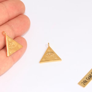 Puede incluir: Colgantes triangulares chapados en oro con un símbolo de ojo y una pequeña etiqueta rectangular que dice "24k Gold Plated". Los colgantes son triangulares con un lazo en la parte superior para colgar. El símbolo del ojo está en el centro del triángulo.