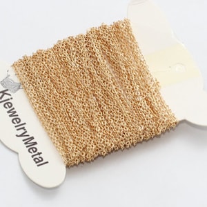 May include: A spool of fine gold-coloured chain, wound on a white card. The card displays the text "KjewelryMetal" in black. The chain is composed of small, interconnected links, creating a textured surface. Suitable for jewellery making.