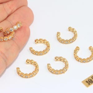 May include: Gold-plated ear cuffs with clear, sparkling stones. The ear cuffs are arranged on a white surface, with a small rectangular tag that reads "24k Gold Plated". The jewelry is designed to be worn on the ear.