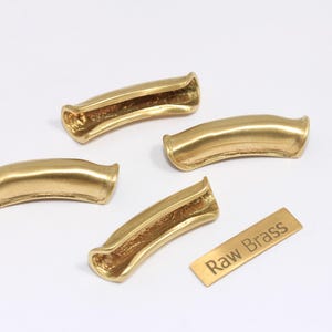 Peut inclure: Quatre poignées de tiroir en laiton brut avec un design incurvé et une étiquette rectangulaire en laiton qui indique "Raw Brass". Les poignées ont un intérieur creux et une finition lisse et polie.