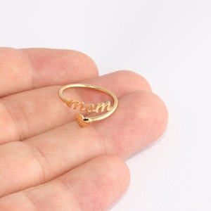 May include: A gold-coloured ring with the word "mom" in a script font. The ring has a heart-shaped accent. The ring is open and adjustable.