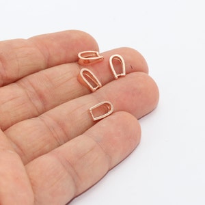 May include: Four rose gold-colored pendant bails. Each bail has a U-shaped design with a flat base, perfect for attaching to necklaces or other jewelry. The bails are small and delicate, ideal for a variety of jewelry projects.