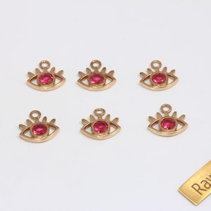 May include: Six gold-colored brass eye charms with a red center stone. Each charm has a loop at the top for stringing. A small brass rectangle with the text "Raw Brass" is also in the image.