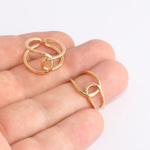 Puede incluir: Dos anillos dorados con un diseño abierto. Los anillos tienen una forma única e entrelazada, con un acabado liso y pulido. Los anillos se muestran sobre un fondo claro.