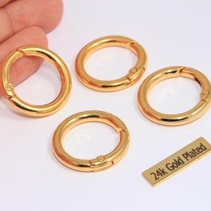 May include: Four gold-plated round spring clasp rings and a small rectangular tag with the text "24k Gold Plated". The rings are open and ready to use. The items are on a white surface.