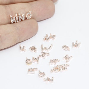 May include: Rose gold-colored letter charms spelling "KIN" and a charm with a small circle. The charms are encrusted with small, clear crystals. Additional letter charms are scattered around the main charms.