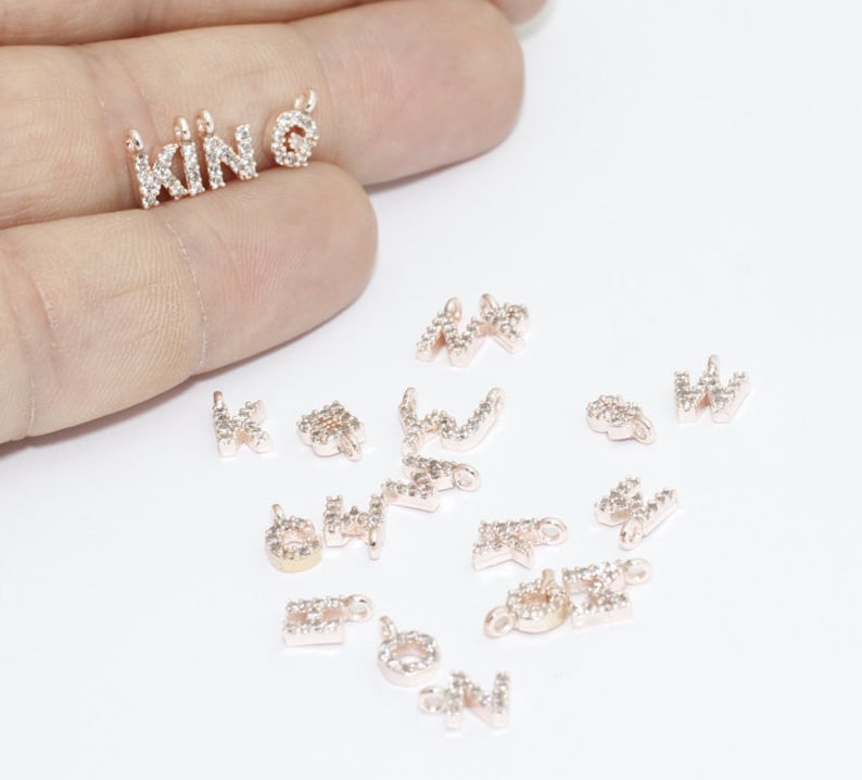 May include: Rose gold-colored letter charms spelling "KIN" and a charm with a small circle. The charms are encrusted with small, clear crystals. Additional letter charms are scattered around the main charms.