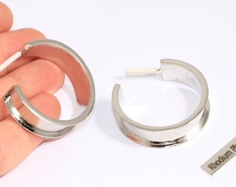 May include: A pair of silver-colored hoop earrings with a polished finish. The earrings have a thick, rounded design and a post closure. A small rectangular tag with the text "Rhodium Plated" is also visible.