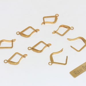May include: A collection of gold-colored earring clasps and a small brass tag with the text "Raw Brass". The clasps are in various shapes, including squares and curved designs. The clasps are made of metal and are on a white background.