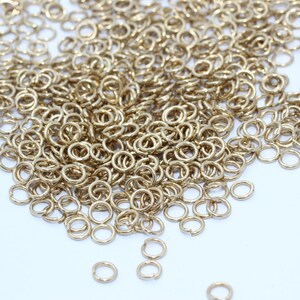 Puede incluir: Una colecci&oacute;n de peque&ntilde;os anillos de salto dorados esparcidos sobre una superficie blanca. Los anillos son circulares con una peque&ntilde;a abertura, lo que sugiere que son para la fabricaci&oacute;n de joyas o manualidades. La iluminaci&oacute;n resalta el brillo met&aacute;lico.