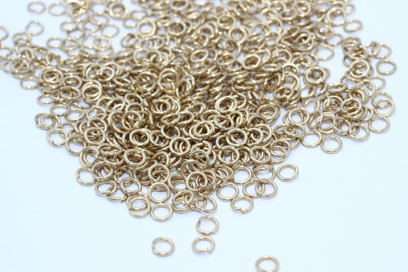 Puede incluir: Una colecci&oacute;n de peque&ntilde;os anillos de salto dorados esparcidos sobre una superficie blanca. Los anillos son circulares con una peque&ntilde;a abertura, lo que sugiere que son para la fabricaci&oacute;n de joyas o manualidades. La iluminaci&oacute;n resalta el brillo met&aacute;lico.