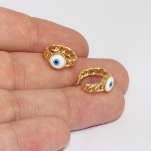 May include: Two gold ear cuffs. Each cuff has a chain design and a circular charm with a white background and a blue eye. The cuffs are small and appear to be made of metal.