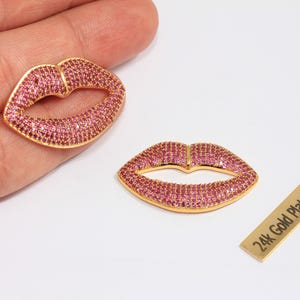 May include: Two gold-plated lip-shaped charms encrusted with pink gemstones. The charms are on a white surface. A small rectangular tag with the text "24k Gold Plated" is also present.