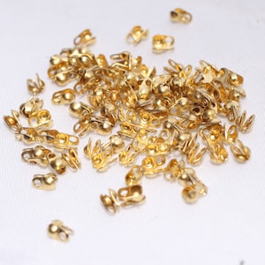 May include: A collection of small, gold-colored crimp end findings, scattered on a white surface. These jewelry-making components have a rounded shape with multiple holes and slots for attaching to cords or ribbons. The findings are designed to secure and finish the ends of jewelry.