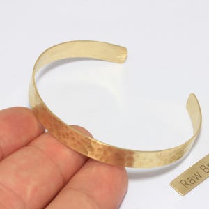 May include: A hammered brass cuff bracelet with a textured surface. The open-ended bracelet is a light gold color and is shown next to a small brass tag that reads "Raw Brass".